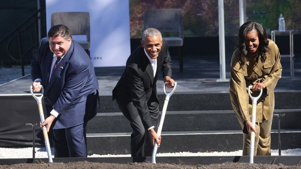 Illinois governor JB Pritzker digging dirt at the site in a ceremonial groundbreaking ceremony with Barack and Michelle Obama.