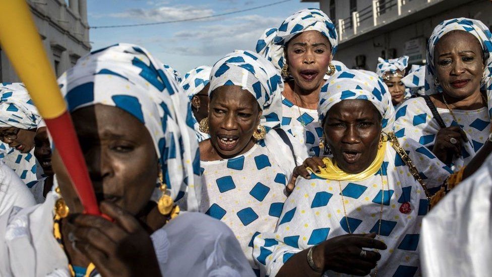 Women dressed in a patterned white a blue outfit with horn blowers. They are celebrating - July
