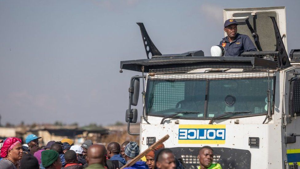 A South African Police Service (SAPS) officer can be seen on top of a Nyala (armoured vehicle) next to residents protesting against illegal mining and rising crime in the area in Kagiso on August 4, 2022.