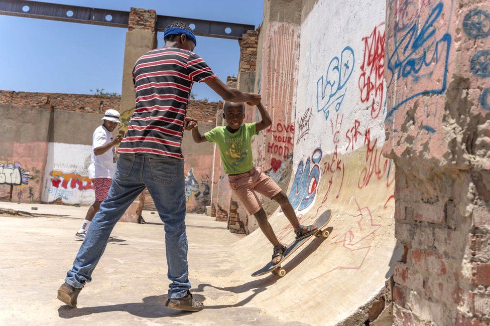 A man holds a child's hands as he skateboards down a ramp.
