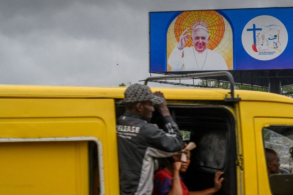 A billboard shows an image of Pope Francis in Kinshasa as passengers travel on a minibus past it.