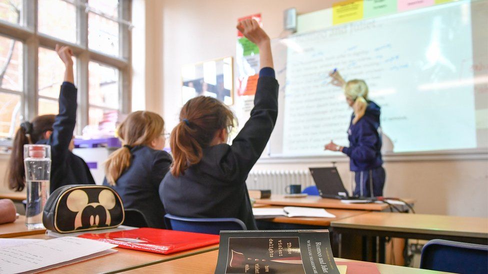 Schoolgirls are seen raising their hands while sat on the front row of a classroom. In front of them, a teacher writes on a whiteboard.