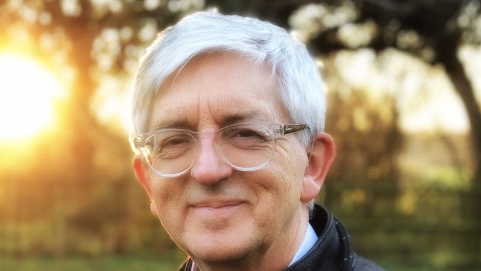 Stephen Sizer, wearing white glasses and with grey hair, smiles looking into the camera with a sunset behind him.