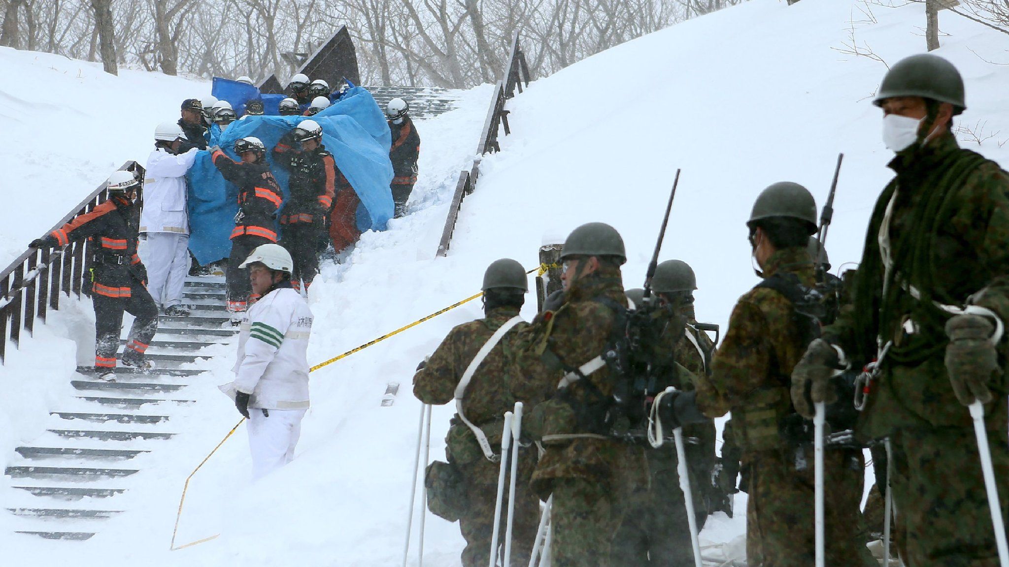 Firefighters carry a survivor they rescued from the site of an avalanche in Nasu town, Tochigi prefecture on 27 March 2017, while Self Defense Force personnel look on