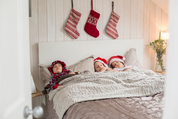 Three children in a double bed, with Christmas stockings hanging on the wall above their heads.